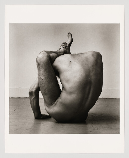 A naked person sits on the floor contorted, holding one foot behind their head.
