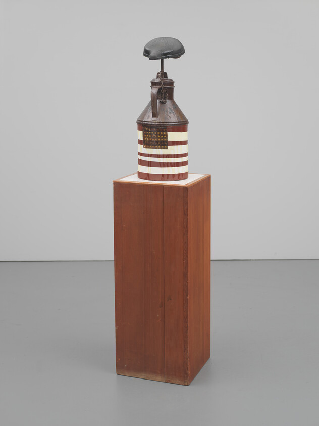 A painted metal can with an American flag motif topped by a dark helmet on a wood pedestal.