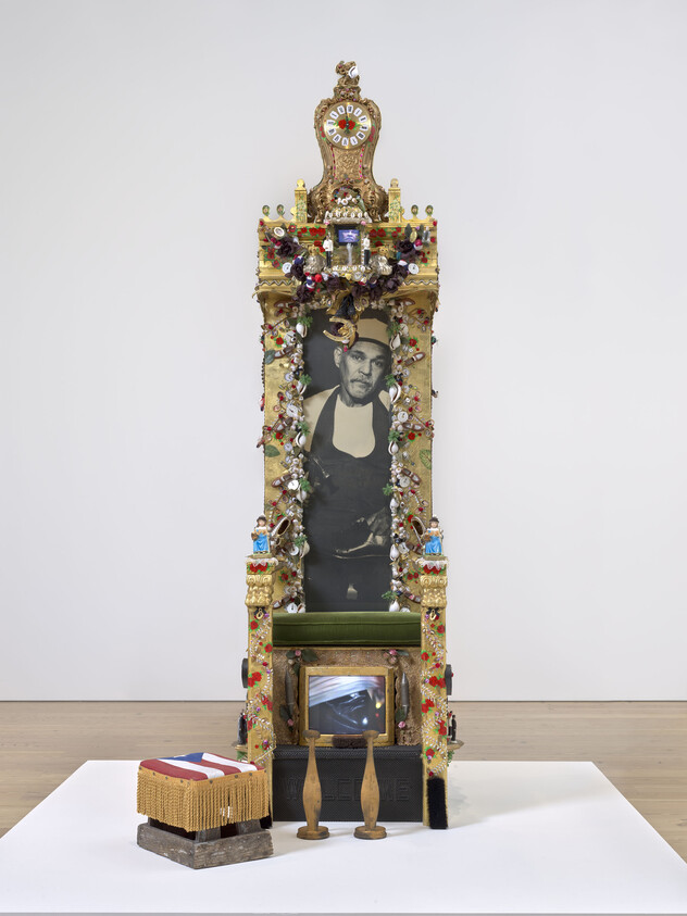 An elaborate gold throne covered in tiny trinkets with small TV monitors under the seat and at the crest. Covering the seatback, a photo of a Puerto Rican man in an apron and cap. Beside the throne, a footstool with a Puerto Rican flag and lined with fringe&nbsp;