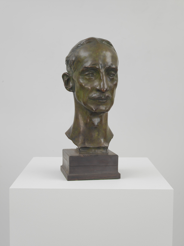 A bronze bust of a man with a solemn expression, displayed on a wooden pedestal atop a white plinth against a plain background.