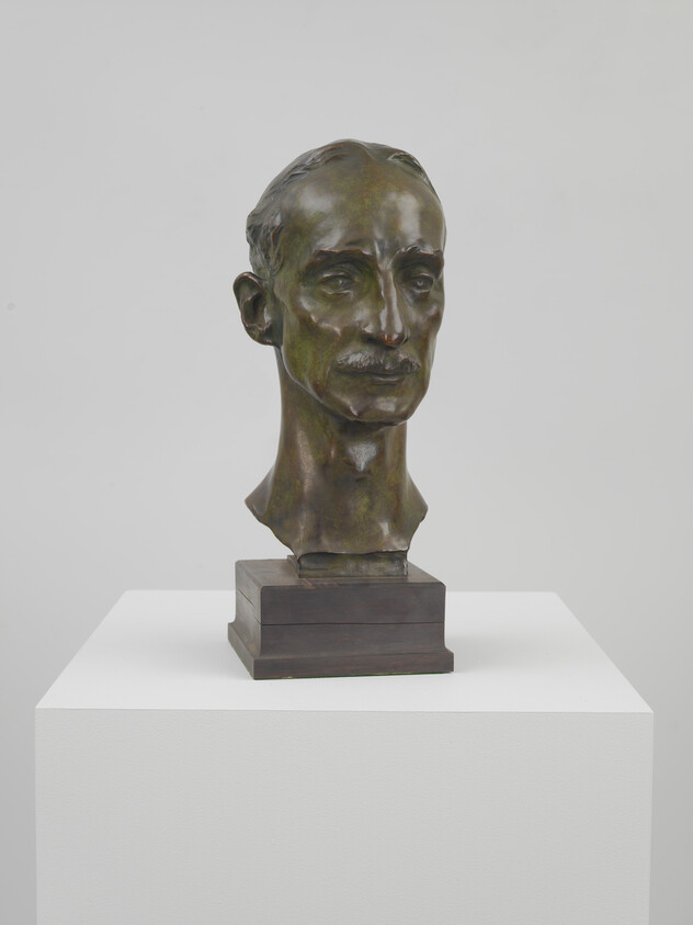 Bronze bust of a middle-aged man with a thin mustache displayed on a white pedestal.