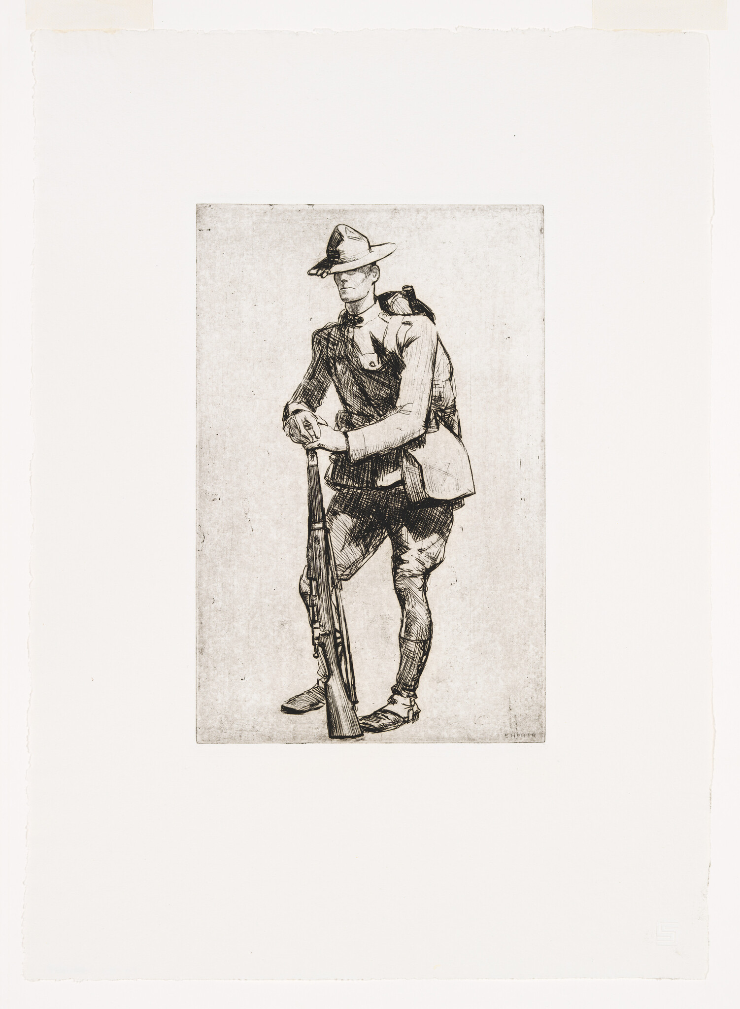 A uniformed soldier stands leaning on his rifle while wearing a wide-brimmed hat and pack.