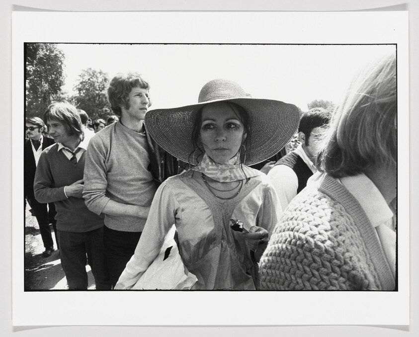 A woman wearing a large sun hat stands in a crowded outdoor line holding a small object.