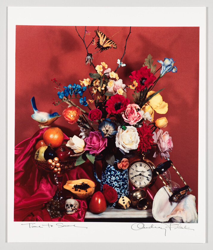 A vibrant floral arrangement in a patterned vase surrounded by a clock, hourglass, fruit, and a small skull.