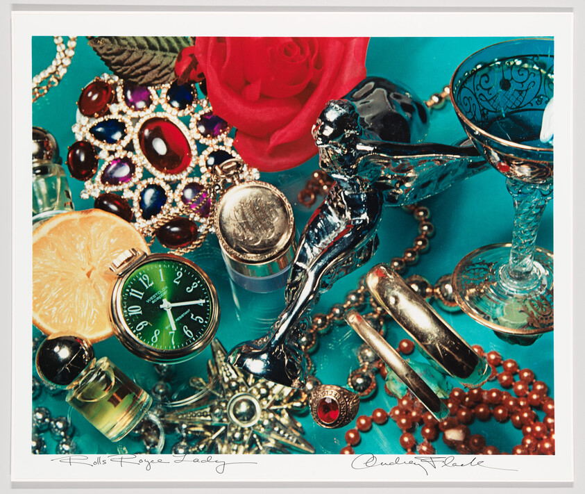 A collection of vintage jewelry, a silver hood ornament, a green watch, and a blue glass on teal.