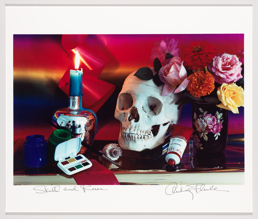 A human skull surrounded by colorful flowers, a lit candle, and makeup items.