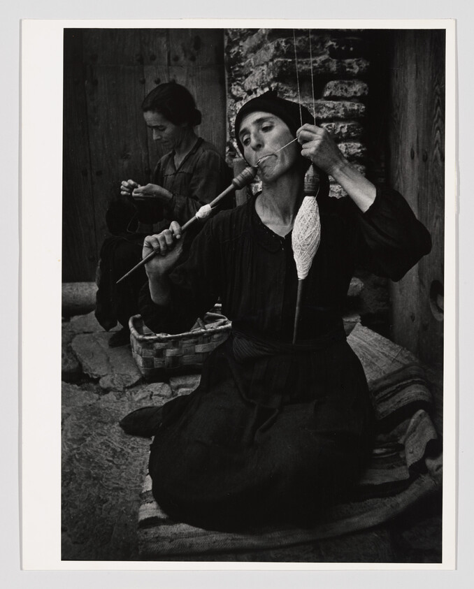 A seated woman uses a spindle and her mouth to spin yarn while another knits behind her.