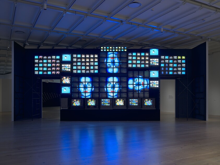 A large wall of televisions displays blue-toned faces in a dark gallery room.