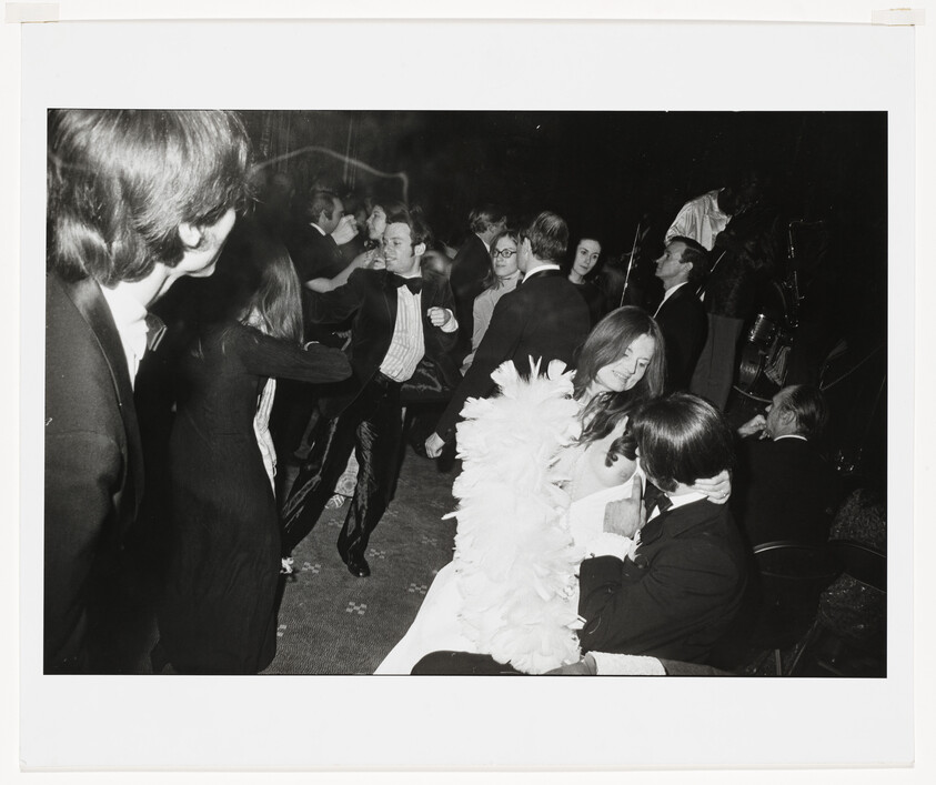 Young woman wearing a feather boa sits on a man's lap while people dance around them.