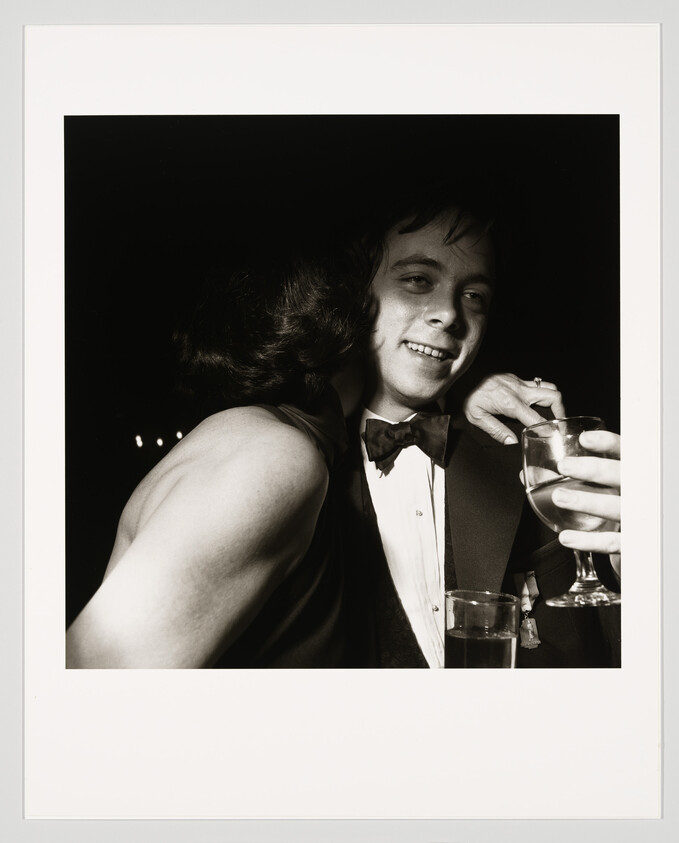 A young man in a tuxedo smiles as a woman hugs him while he holds a drink.
