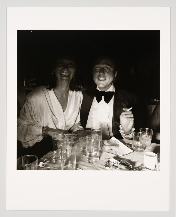 A smiling couple sits at a cluttered table while a man in a tuxedo holds a cigarette.