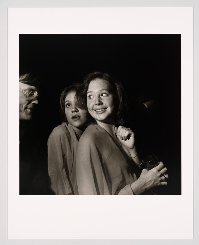 Two young women huddle closely at a dim party as one smiles and holds a drink.