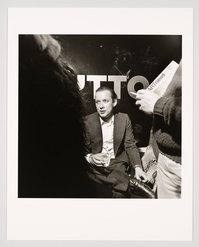 A man in a suit sits holding a drink while people stand and talk around him.