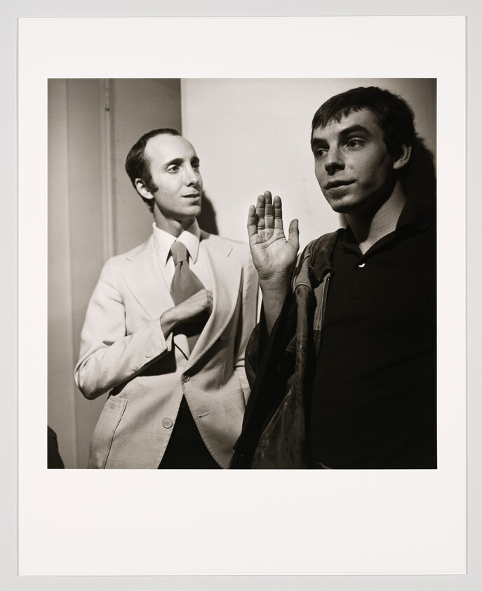 Two men stand by a wall, one in a suit adjusting his tie while the other raises his hand.