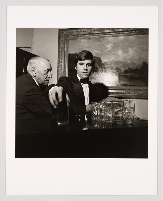 Young man in a tuxedo reaches for a glass at a bar while an older man watches.