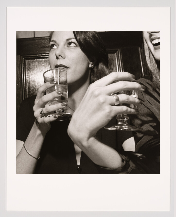 Two women hold drinks at a bar, one looking up while the other laughs.