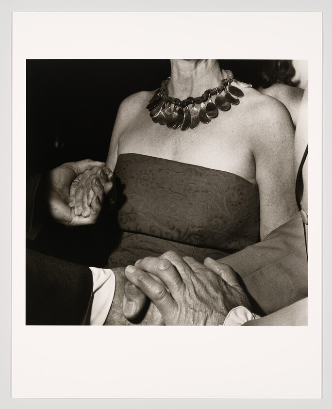 A woman in a strapless dress and bold necklace has several hands resting on her arms and hands.