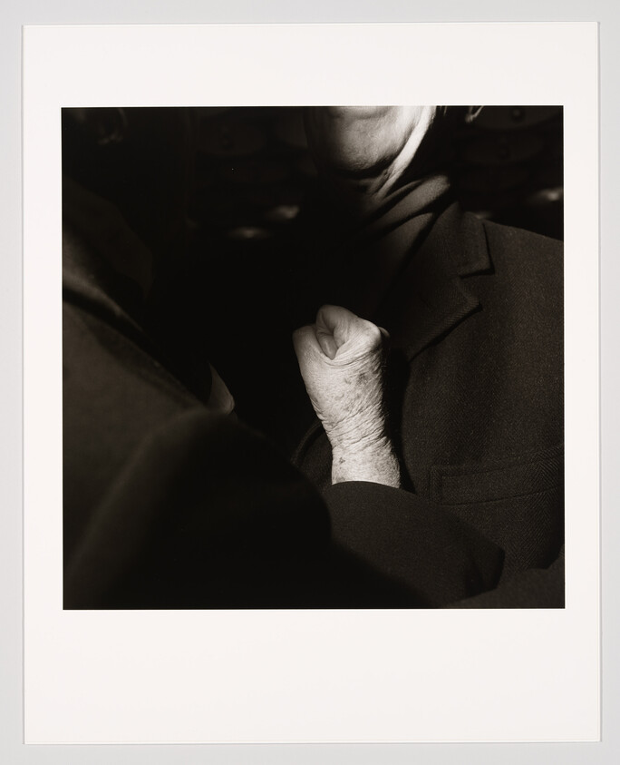 An elderly hand clenches another person's lapel near the chest in a dimly lit close-up.