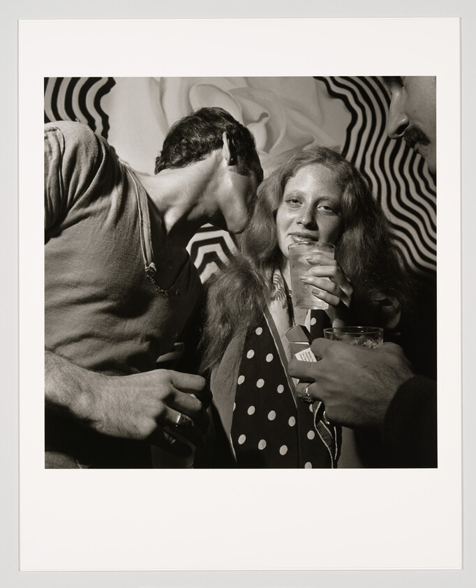 Young woman holding a drink while a man whispers in her ear at a crowded party.