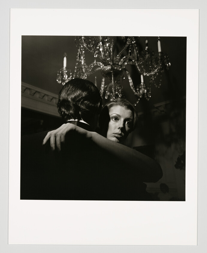 Woman embraces a man while dancing beneath a crystal chandelier, her face looking distant.
