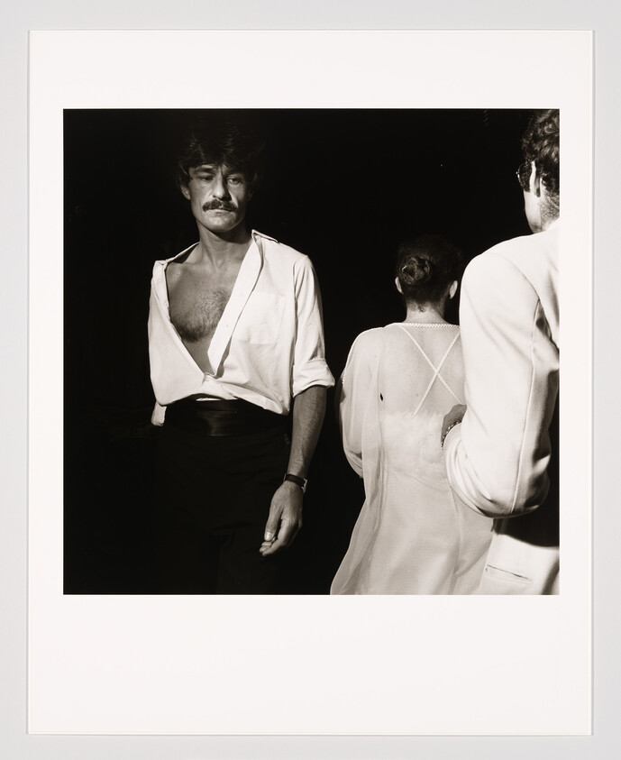 Man with open white shirt and mustache stands beside two people dressed in light clothing.