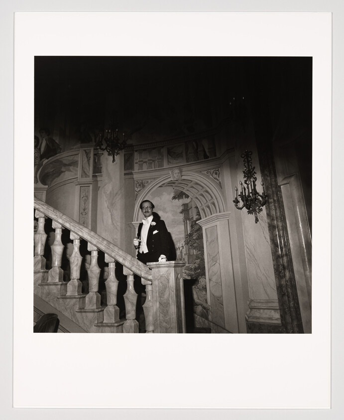 A man in a tuxedo stands on an ornate marble staircase holding a drink.