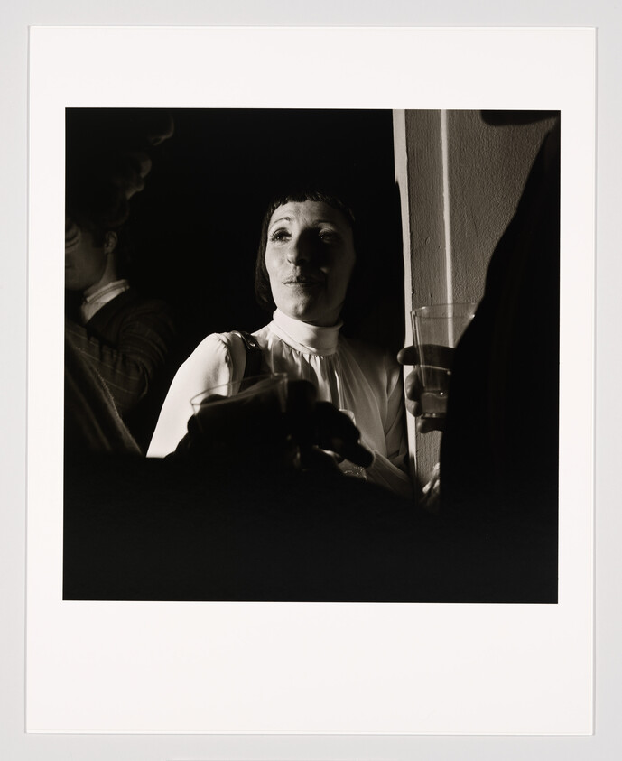 Woman in a white blouse stands by a wall holding a drink while talking with others.