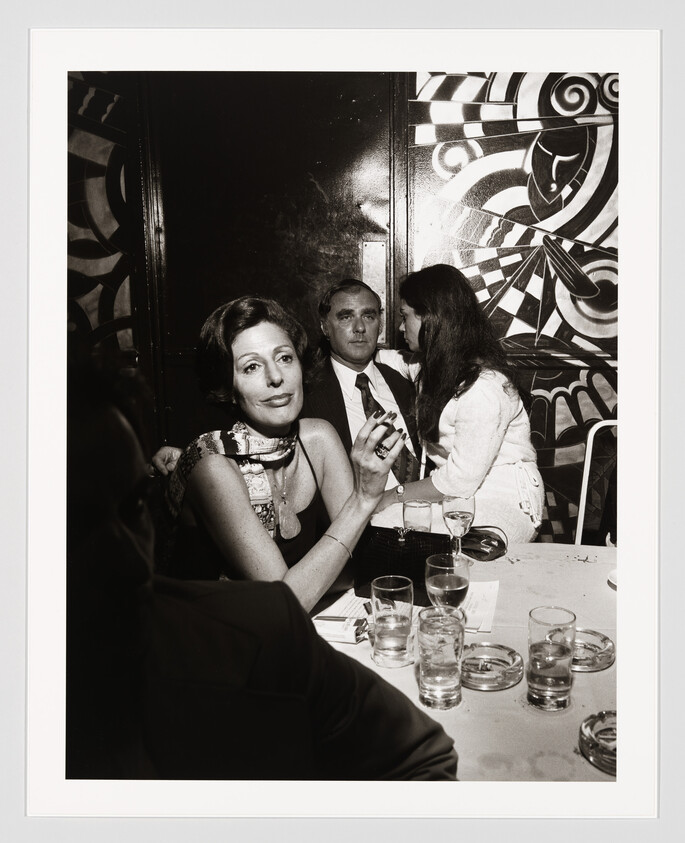 Woman holding a cigarette at a busy table while two people sit and talk behind her.