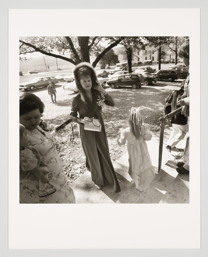 A woman in a fur hat and long dress walks up steps with a young girl and papers.