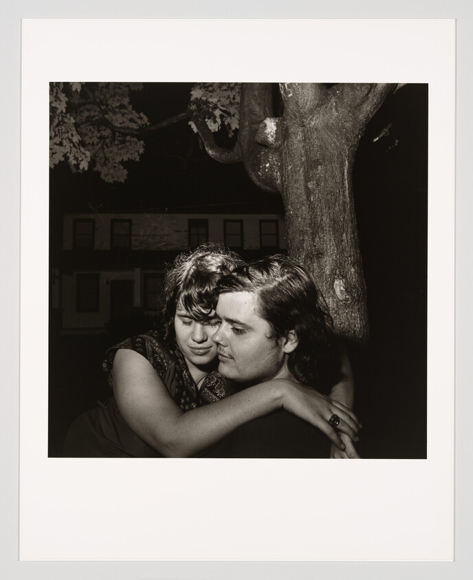 A young couple embraces closely by a tree at night, eyes closed and thoughtful.