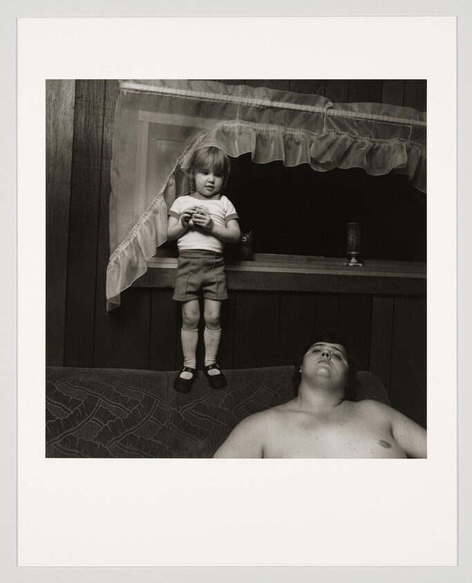 Young child stands on couch back near window holding a toy while a shirtless adult reclines below.