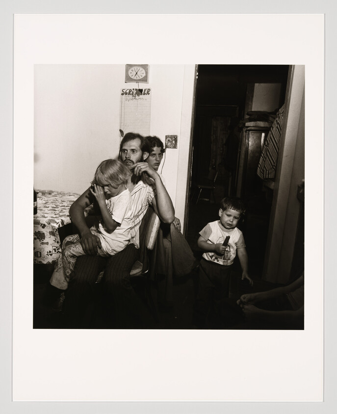 A tired father sits with two young children, one crying and one holding a toy.