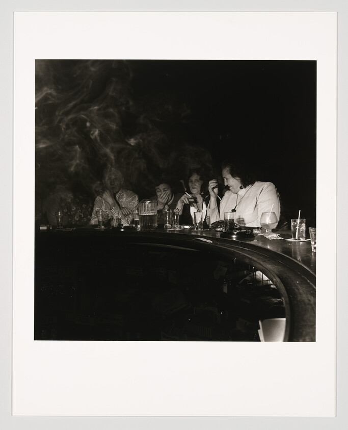 Four people sit at a curved bar smoking and talking over drinks late at night.