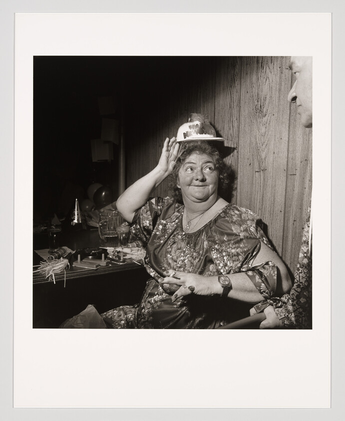 A black and white photograph of a smiling woman seated at a party, wearing a festive hat with "Happy New Year" written on it. She is raising her hand in a wave or greeting, and there are party decorations on the table in front of her. A partial view of another person is visible to the right of the frame.