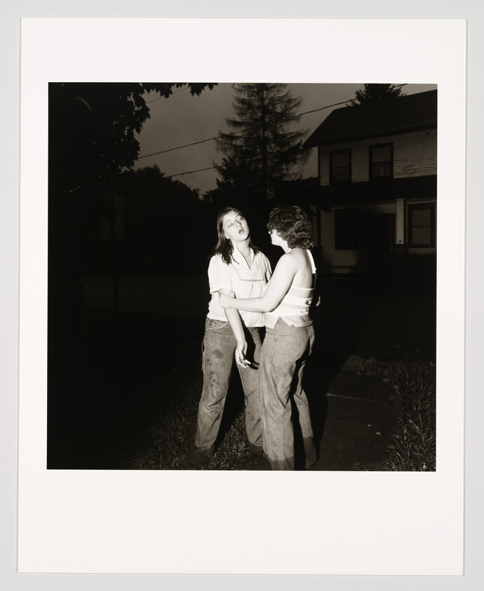 Two teenagers stand on a dark suburban sidewalk at night while one supports the other.
