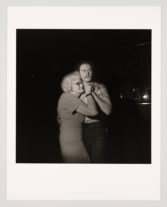 An elderly woman embraces a younger man as they stand closely together in dim light.
