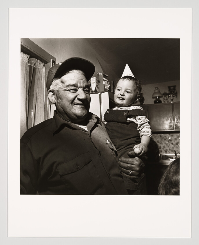 An older man holds a smiling baby wearing a small paper party hat in a kitchen.