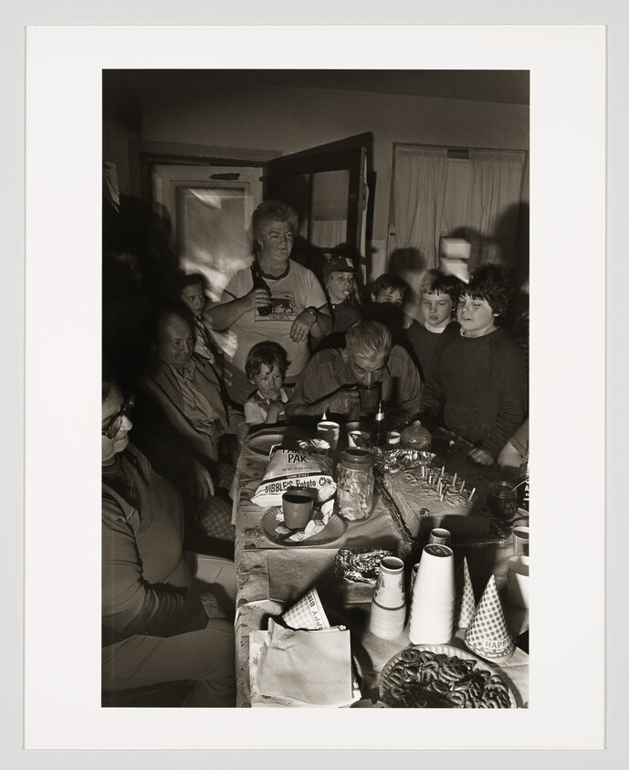 A man leans over a birthday cake surrounded by family watching him blow out candles.