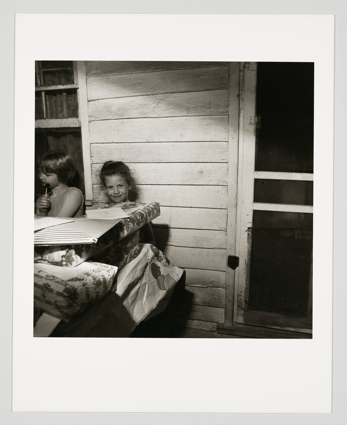 Two children sit by a wooden wall with wrapped presents, one girl smiling at the camera.