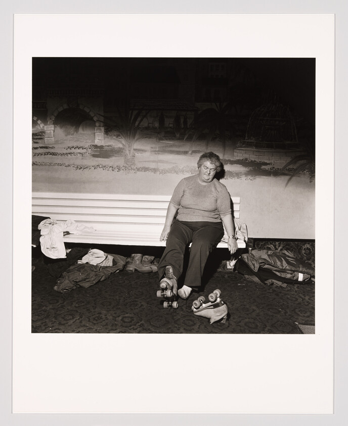 A woman sits on a bench putting on roller skates with extra skates and clothes nearby.