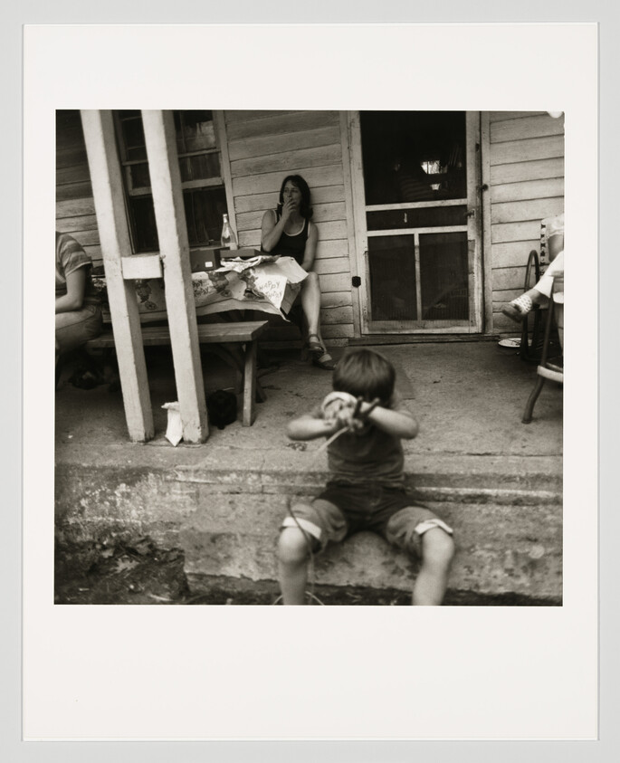 A young boy sits on concrete steps covering his face while a woman smokes on the porch.