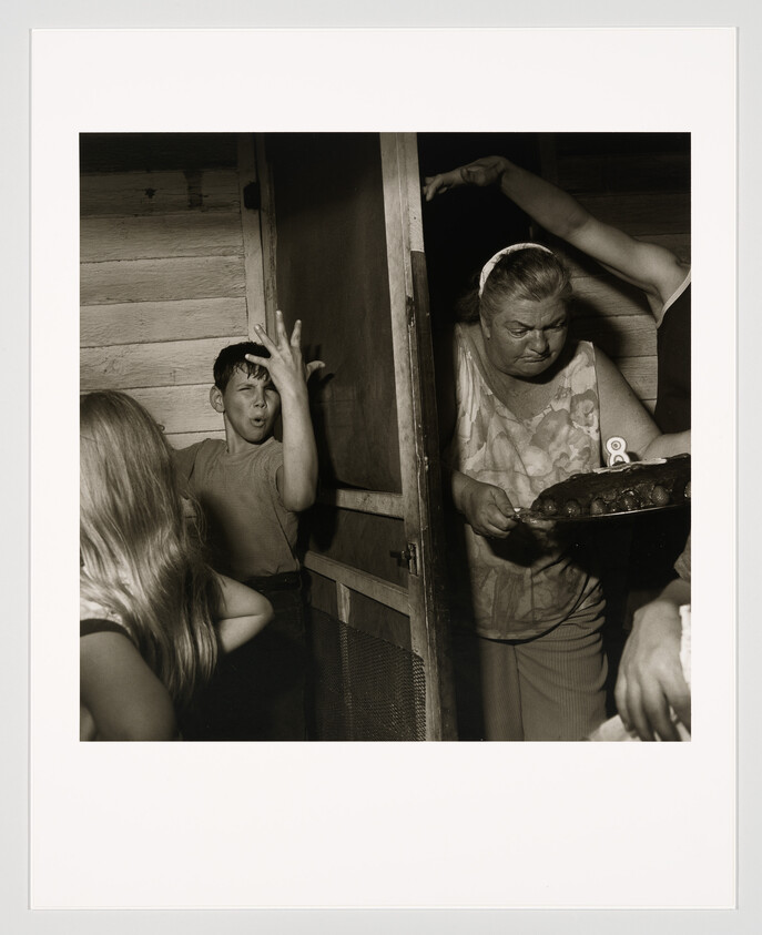 An older woman carries a small cake while children react excitedly near a doorway.