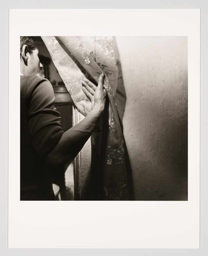 A black and white photograph of a person from behind, partially obscured, looking out a window through a sheer curtain with floral patterns. The individual appears to be holding the curtain aside with one hand, possibly peering outside. The image conveys a sense of curiosity or anticipation.