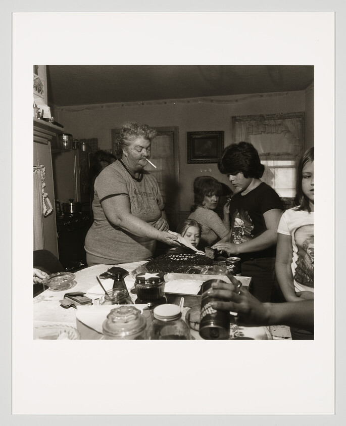 A black and white photograph capturing a family gathering in a kitchen, where an older woman with a cigarette in her mouth is cutting a cake. Several individuals, including children and adults, are gathered around the table filled with various items like soda cans, condiments, and kitchenware. The atmosphere appears casual and homey.