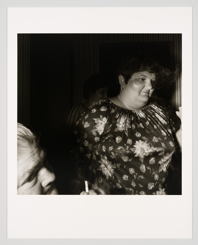 A woman in a floral blouse stands smoking while another person holds a lit cigarette nearby.