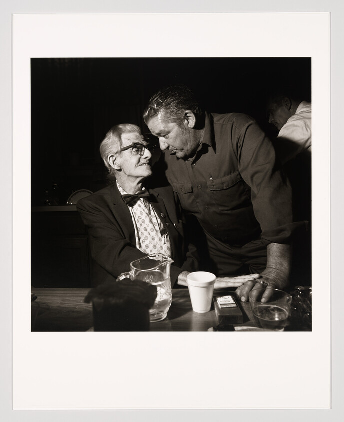 An older man in a bow tie sits as another man leans in to speak across the table.