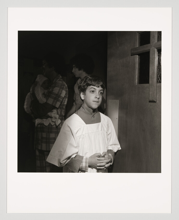 A boy in a white altar robe stands with hands clasped near a church door cross.