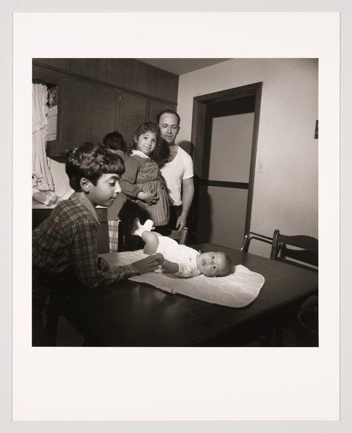 Father and children gathered around a table while an older boy tends to a baby on a changing pad.
