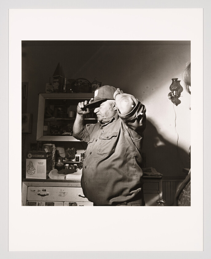 An older man stands in a cluttered kitchen putting on a baseball cap.