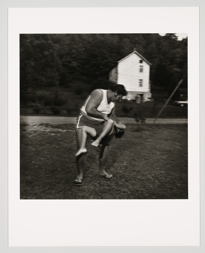 A man in shorts playfully swings a child upside down on a grassy yard near a house.